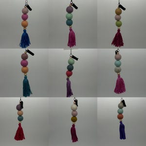 May include: Nine colorful felt ball and tassel keychains with a clip on the top. The keychains are in various colors including pink, blue, green, and beige.