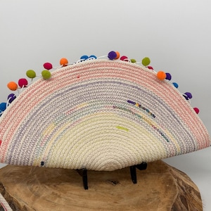 May include: A half-circle shaped clutch with a rainbow design. The clutch is made of woven rope and features a variety of colors, including pink, orange, purple, and white. It is decorated with colorful pom-poms along the top edge and has a white rope strap.