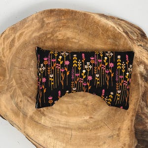 May include: A black eye pillow with a floral pattern in pink, yellow, and white. The pillow is rectangular with a curved cutout on one side. The pillow is placed on a wooden surface.