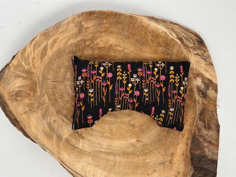 May include: A black eye pillow with a floral pattern in pink, yellow, and white. The pillow is rectangular with a curved cutout on one side. The pillow is placed on a wooden surface.