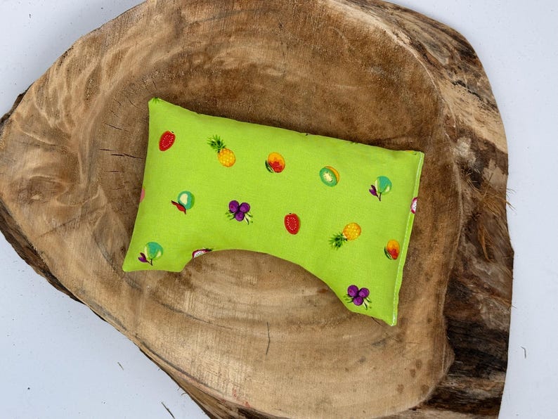 May include: A small, lime green pillow with a fruit pattern, including strawberries, pineapples, grapes, and kiwi. The pillow is rectangular with a curved indentation on one side. The pillow is on a wooden surface.