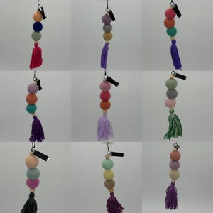May include: A set of ten colorful felt ball car charms with a tassel. Each charm has three felt balls and a clip to attach to a car's rearview mirror. The colors include pink, blue, green, purple, and orange.