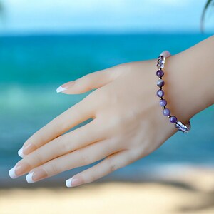 May include: A purple and white gemstone bracelet with gold accents. The bracelet is on a hand with white nail polish.