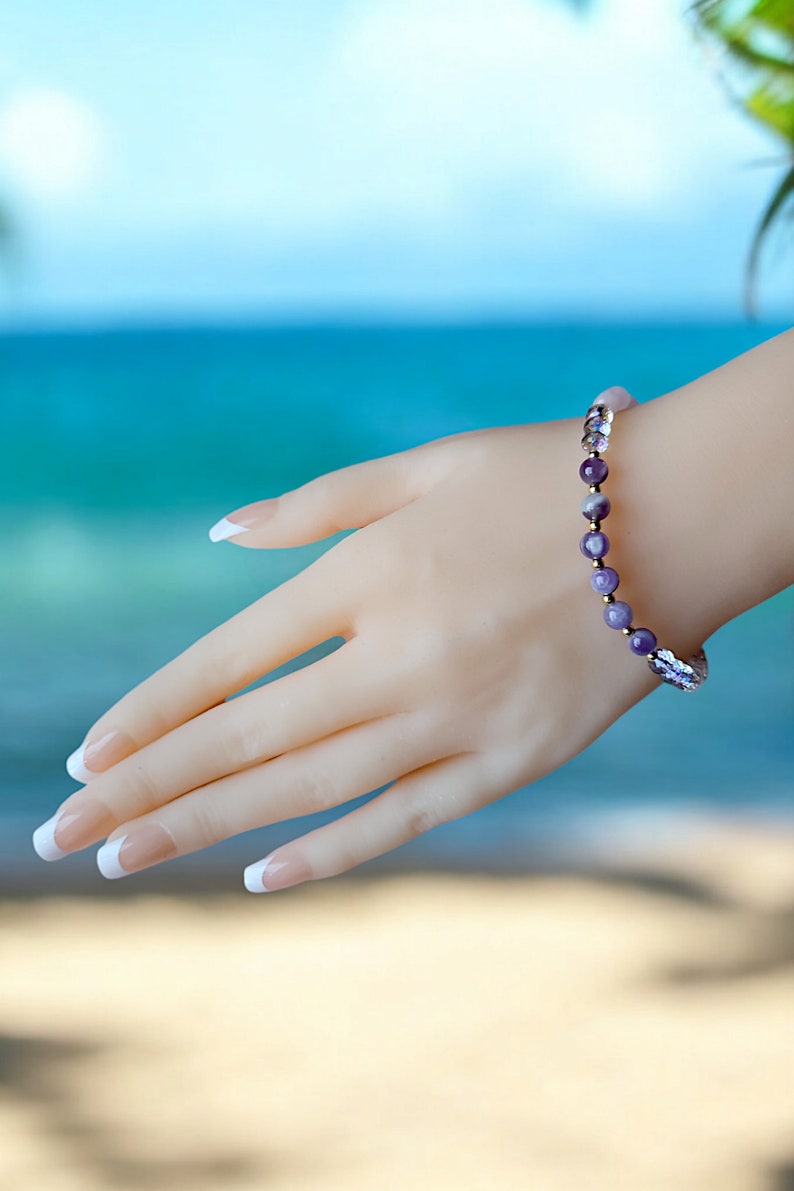 May include: A purple and white gemstone bracelet with gold accents. The bracelet is on a hand with white nail polish.
