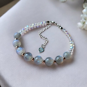 May include: A silver chain bracelet with a heart charm and a row of iridescent grey beads. The bracelet is on a white surface.