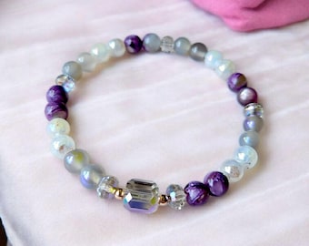 May include: A beaded bracelet featuring a mix of purple, gray, and clear beads. The bracelet has a central rectangular bead with iridescent coloring and is set against a soft pink background.