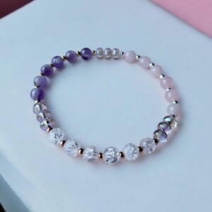 May include: A purple, clear, and pink beaded bracelet with gold accents. The bracelet is made with amethyst, clear quartz, and rose quartz beads.