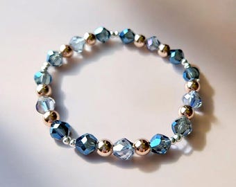 May include: A beaded bracelet featuring faceted blue crystal beads and gold-colored metallic beads. The bracelet is strung on a silver-colored elastic cord. The bracelet is a delicate piece of jewelry.