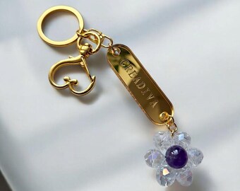 May include: Gold-toned keychain with a heart-shaped clasp and a rectangular tag engraved with "CREADIVA." The keychain features a flower charm made of clear crystals surrounding a purple bead. The background is white.