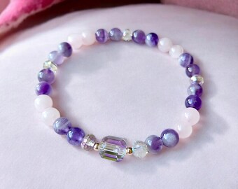 May include: A beaded bracelet featuring alternating purple and pink gemstones. The bracelet includes a central rectangular crystal bead and smaller clear crystal accents. The beads are strung on an elastic band.