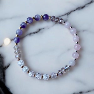 May include: A purple, white, and pink beaded bracelet with gold accents. The bracelet is made with amethyst, clear quartz, and rose quartz beads.