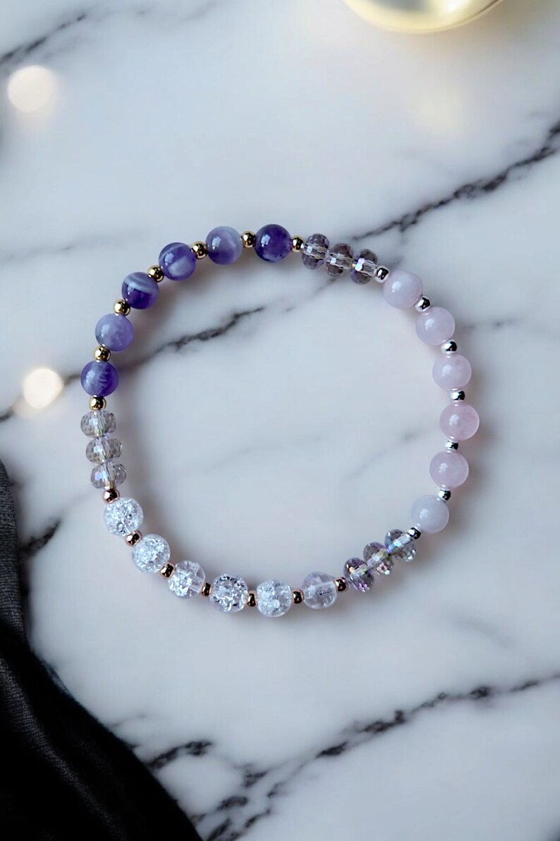 May include: A purple, white, and pink beaded bracelet with gold accents. The bracelet is made with amethyst, clear quartz, and rose quartz beads.
