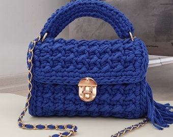 May include: A royal blue, hand-crocheted handbag with a gold-tone chain strap and clasp. The bag features a braided texture, a top handle, and a tassel detail. The bag's dimensions are approximately 10 inches wide, 7 inches high, and 4 inches deep.