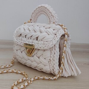 May include: A white crocheted handbag with a gold chain strap and a tassel. The bag has a woven design and a gold clasp.