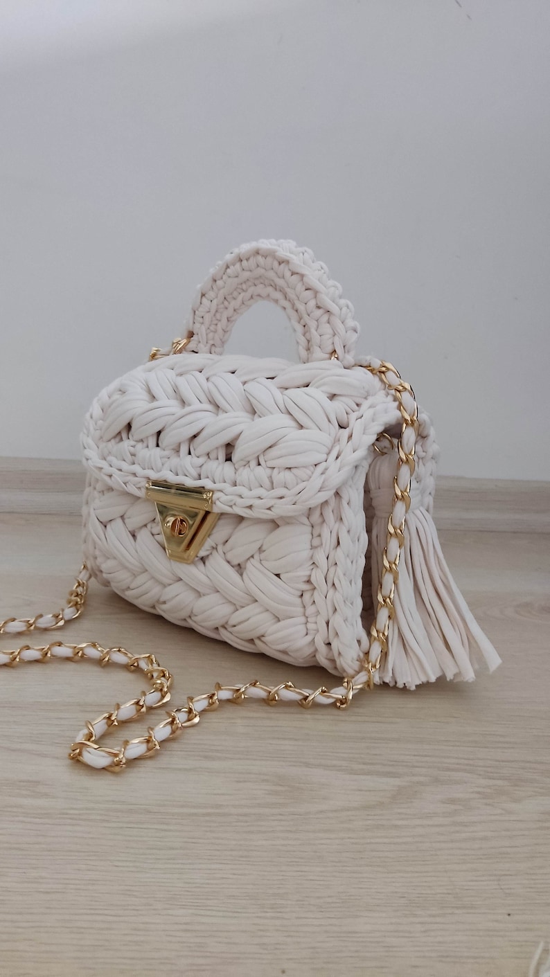 May include: A white crocheted handbag with a gold chain strap and a tassel. The bag has a woven design and a gold clasp.