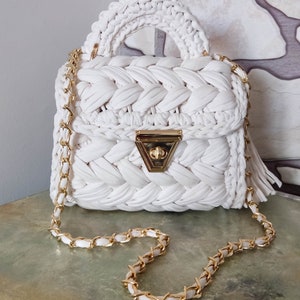May include: A white crocheted handbag with a gold chain strap and a gold clasp. The bag has a braided design and a tassel.