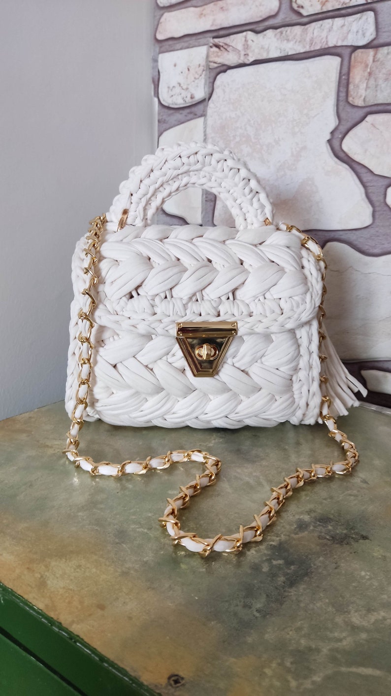 May include: A white crocheted handbag with a gold chain strap and a gold clasp. The bag has a braided design and a tassel.