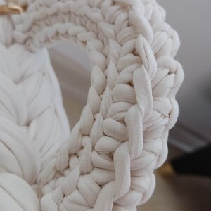 May include: Close-up of a white crocheted bag handle with a gold metal clasp. The handle is made of thick, braided yarn and has a woven texture.