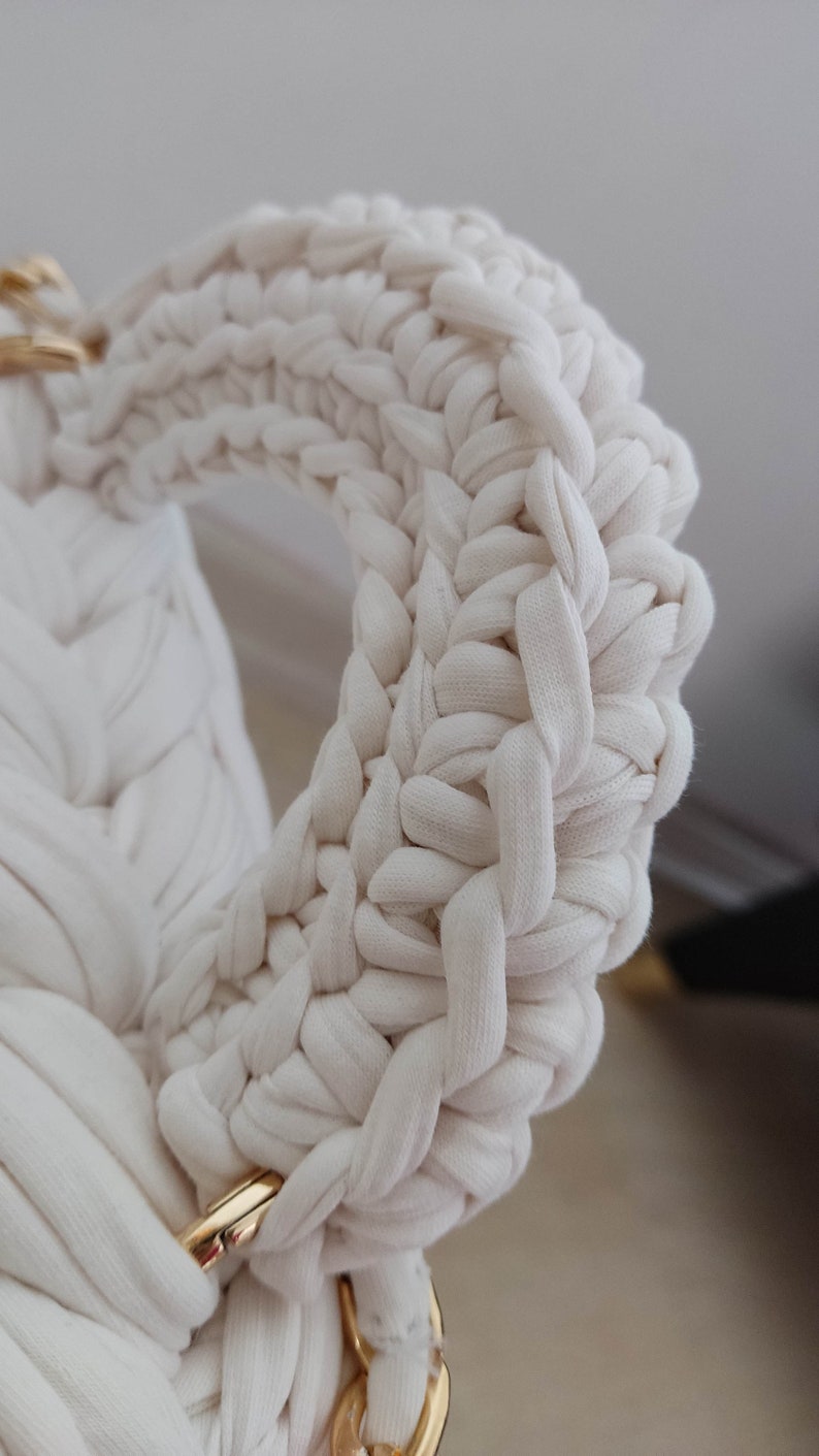 May include: Close-up of a white crocheted bag handle with a gold metal clasp. The handle is made of thick, braided yarn and has a woven texture.