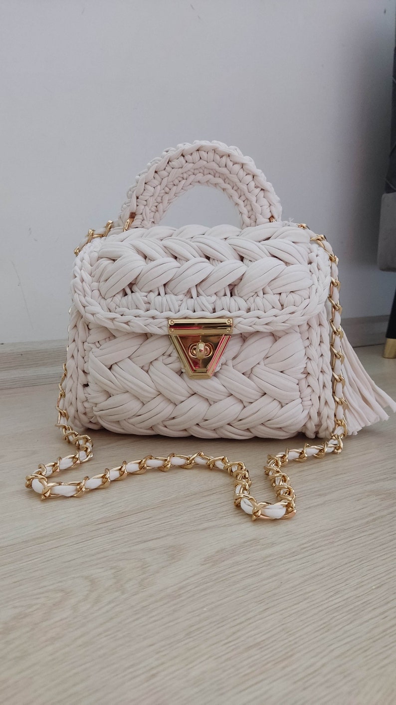 May include: A white crocheted handbag with a gold chain strap and a gold clasp. The bag has a woven texture and a tassel on the strap.
