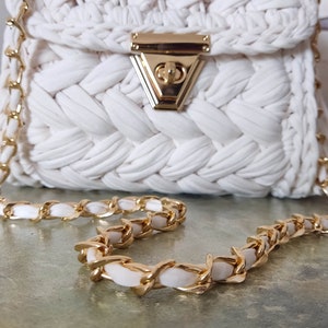 May include: A white crocheted handbag with a gold chain strap and a gold clasp. The bag has a woven texture and a simple design.