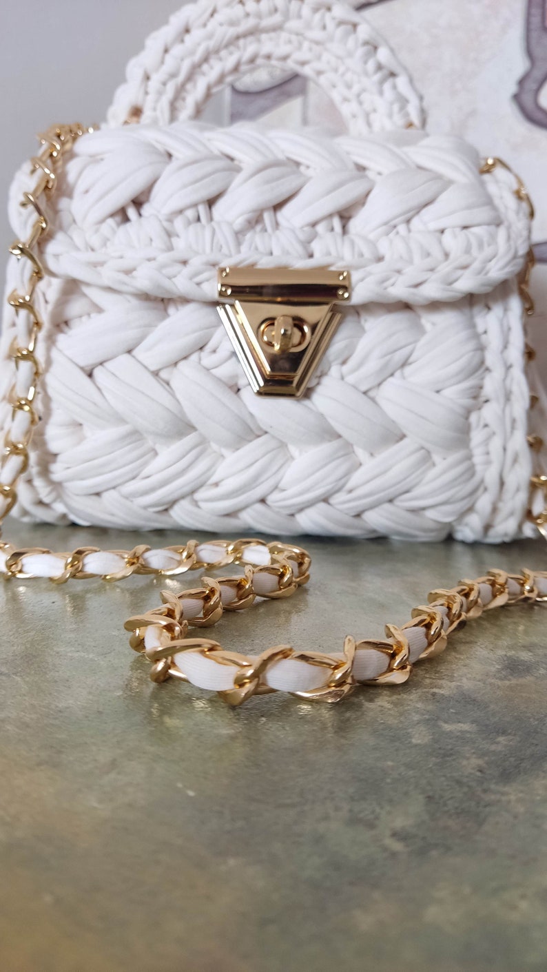 May include: A white crocheted handbag with a gold chain strap and a gold clasp. The bag has a woven texture and a simple design.