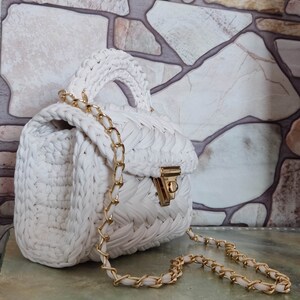 May include: White crocheted handbag with a gold chain strap and a gold clasp. The bag is sitting on a textured surface.