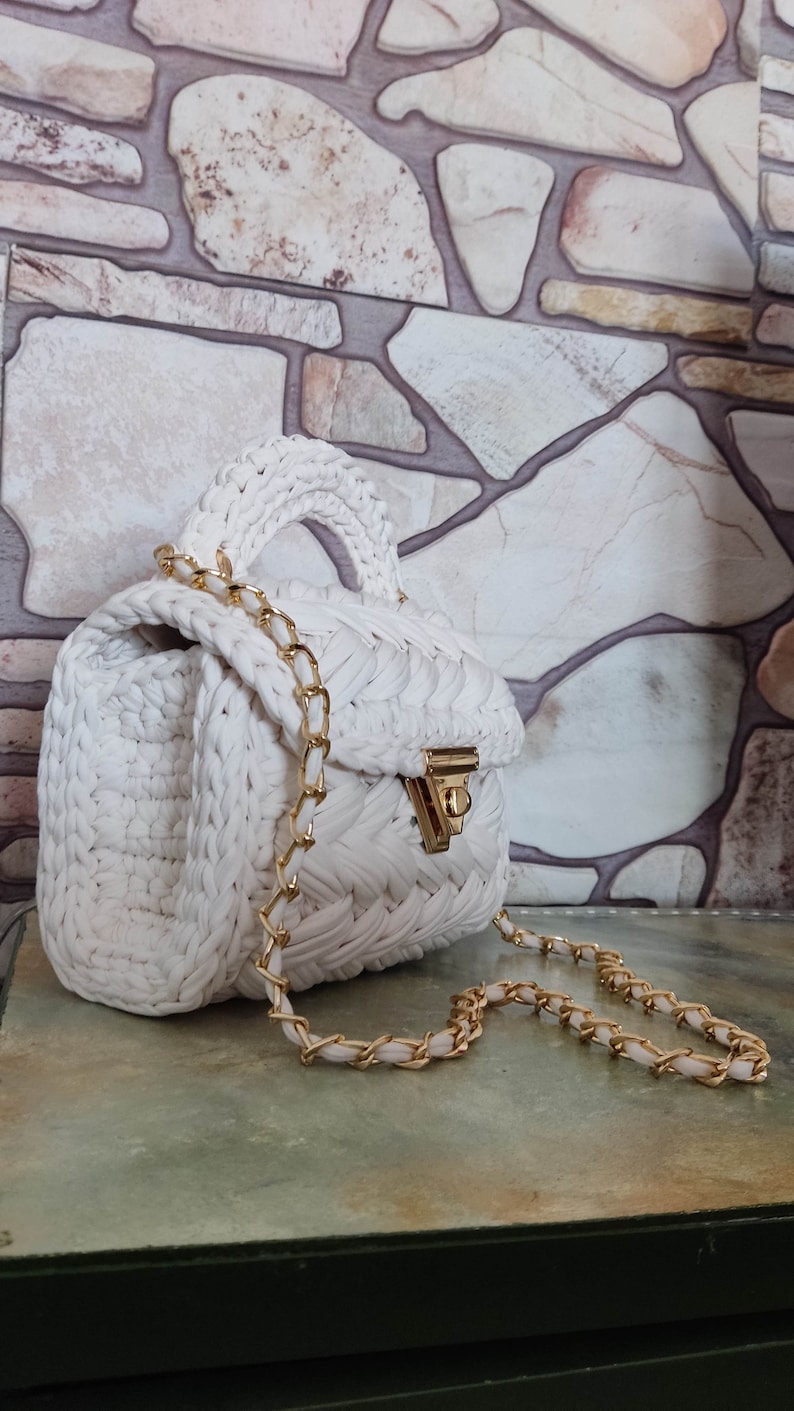 May include: White crocheted handbag with a gold chain strap and a gold clasp. The bag is sitting on a textured surface.
