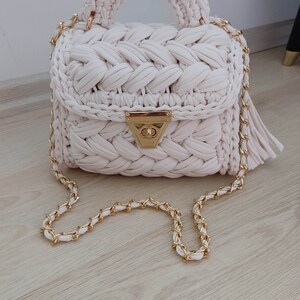 May include: A white crocheted handbag with a gold chain strap and a gold clasp. The bag has a woven design and a top handle.