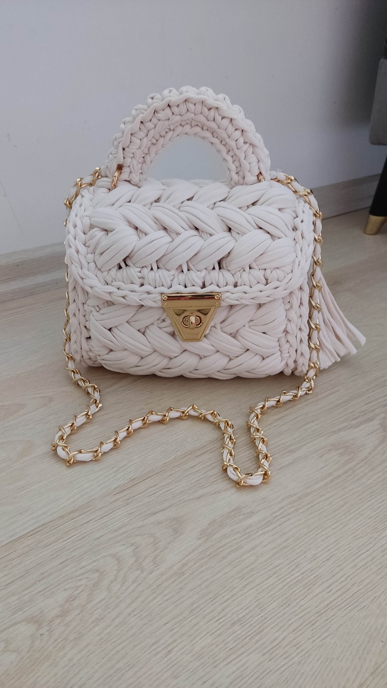 May include: A white crocheted handbag with a gold chain strap and a gold clasp. The bag has a woven design and a top handle.
