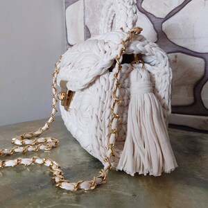 May include: A white crocheted handbag with a gold chain strap and a white tassel. The bag has a unique woven design and a top handle.