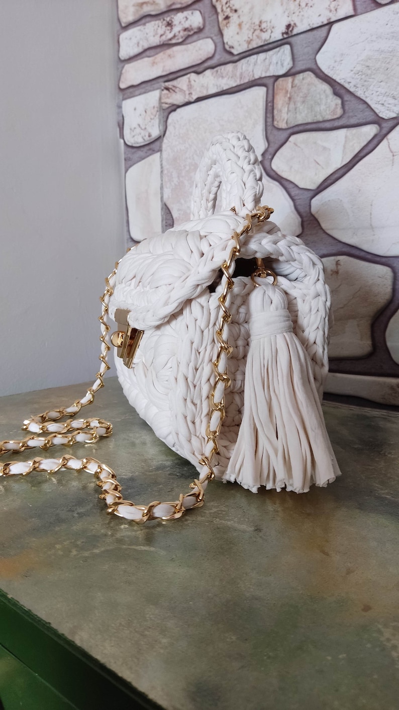 May include: A white crocheted handbag with a gold chain strap and a white tassel. The bag has a unique woven design and a top handle.