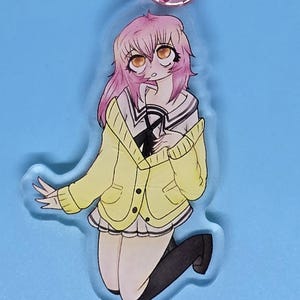 May include: Anime-style acrylic keychain featuring a kneeling girl with pink hair, wearing a yellow cardigan and black knee-high socks. The keychain has a heart-shaped clasp in a rose gold color. The background is a solid light blue.