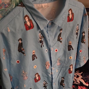 May include: A light blue button-up shirt with a repeating pattern of cartoon characters. The characters are all wearing different outfits and have different expressions. The shirt has a collar and short sleeves.