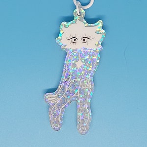 May include: A white and iridescent glitter charm with a cartoon-like figure, featuring a star-shaped clasp. The charm has a whimsical design with a light blue background.