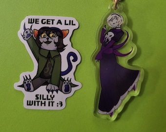 May include: Two acrylic keychains. One features a cartoon cat wearing a green jacket and the text "We get a lil silly with it :3". The other features a cartoon woman in a purple dress and a black hooded cloak.