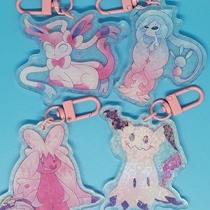 May include: Four clear plastic keychains with pink metal rings. Each keychain features a different Pokemon character: Sylveon, Gardevoir, Mimikyu, and Glaceon. The characters are depicted in a pastel pink and blue color scheme.