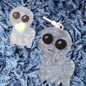 May include: Two translucent white ghost-shaped earrings with black eyes and mouths. The earrings are on a blue fuzzy surface.