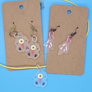 May include: Two pairs of earrings and a necklace featuring translucent, hand-painted resin charms with a whimsical, cartoon-like design. The charms are shaped like a creature with a large eye, a small mouth, and four small feet. The earrings are on brown card stock, and the necklace is on a yellow cord.