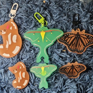 May include: Five keychains featuring different designs: a brown cat, a green moth, a brown butterfly, a brown butterfly, and a brown cat. The keychains are made of a soft, flexible material and have a glossy finish.