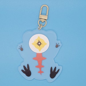 Puede incluir: Un llavero de un personaje de dibujos animados azul y rojo con un ojo grande y pies negros. El llavero est&aacute; unido a un anillo de metal plateado.