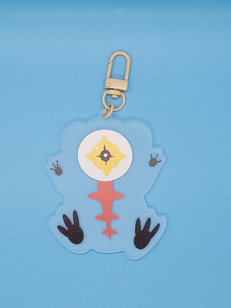 Puede incluir: Un llavero de un personaje de dibujos animados azul y rojo con un ojo grande y pies negros. El llavero est&aacute; unido a un anillo de metal plateado.
