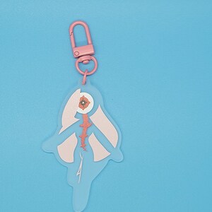 Puede incluir: Llavero rosa con una figura transl&uacute;cida de una persona con un ojo grande y una flor rosa en su interior. La figura tiene un dise&ntilde;o rosa y blanco y una columna vertebral roja.