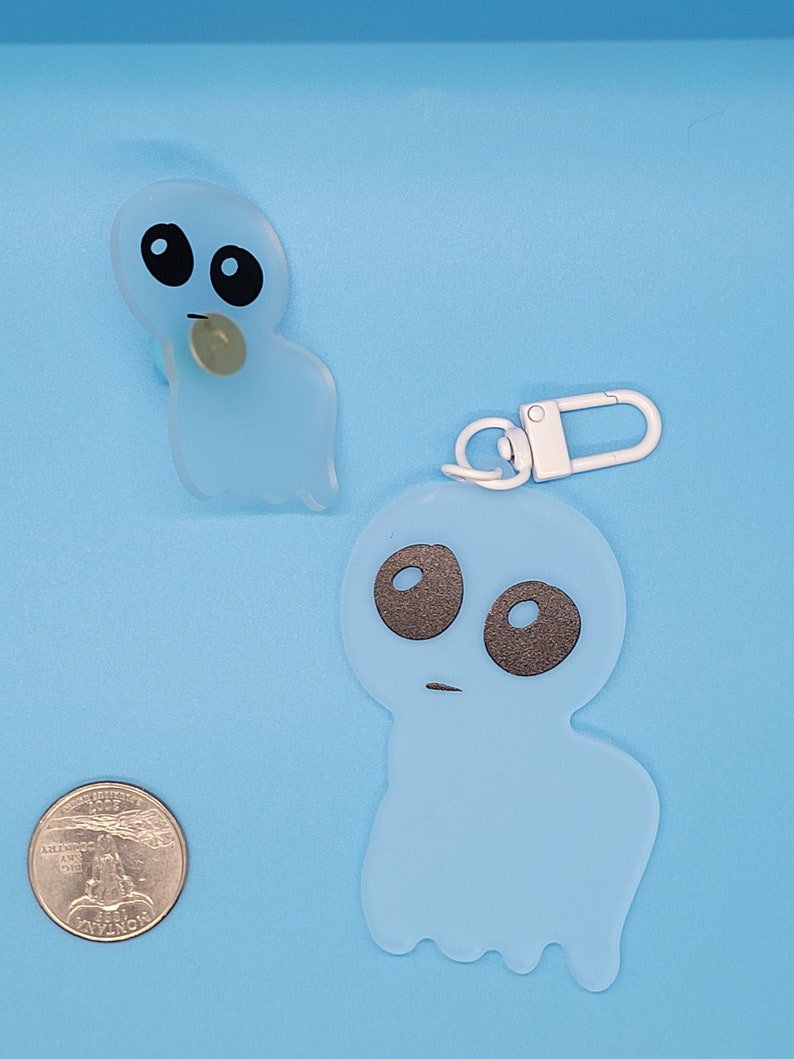 May include: Two blue ghost-shaped keychains with black eyes and a white keychain ring. The larger ghost keychain has a white loop for attaching to a bag or belt.