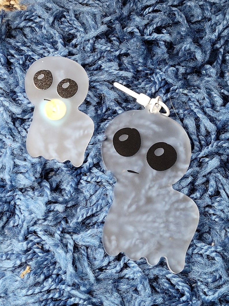 May include: Two translucent white ghost-shaped earrings with black eyes and mouths. The earrings are on a blue fuzzy surface.