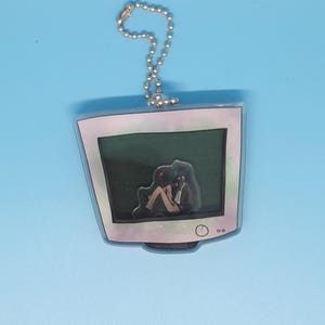 May include: A clear acrylic keyring with a retro computer monitor design. Inside the monitor is a dark scene with a figure. The keyring has a silver ball chain for attachment. The monitor is outlined in white.