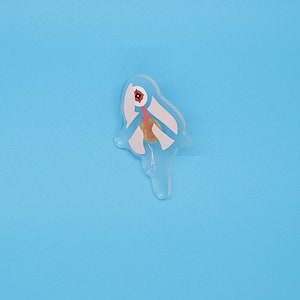 Puede incluir: Un pin de resina blanco, azul y rojo con forma de p&aacute;jaro con un pico largo. El p&aacute;jaro est&aacute; mirando hacia la derecha.