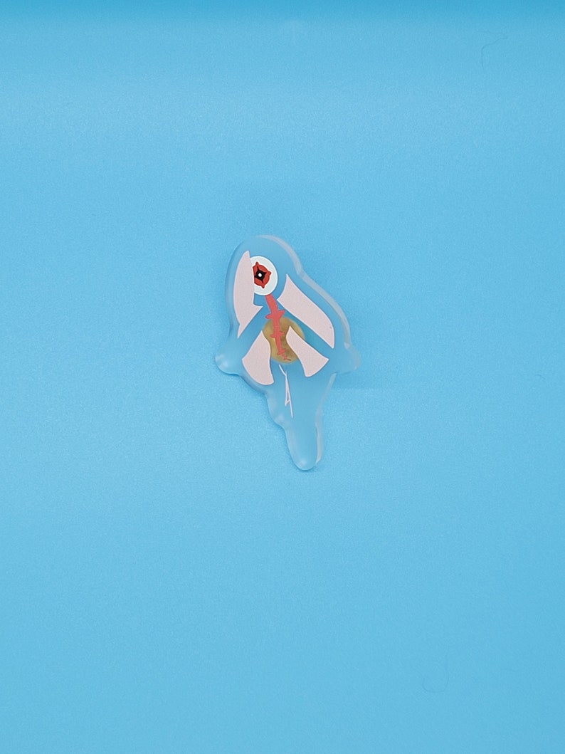 Puede incluir: Un pin de resina blanco, azul y rojo con forma de p&aacute;jaro con un pico largo. El p&aacute;jaro est&aacute; mirando hacia la derecha.