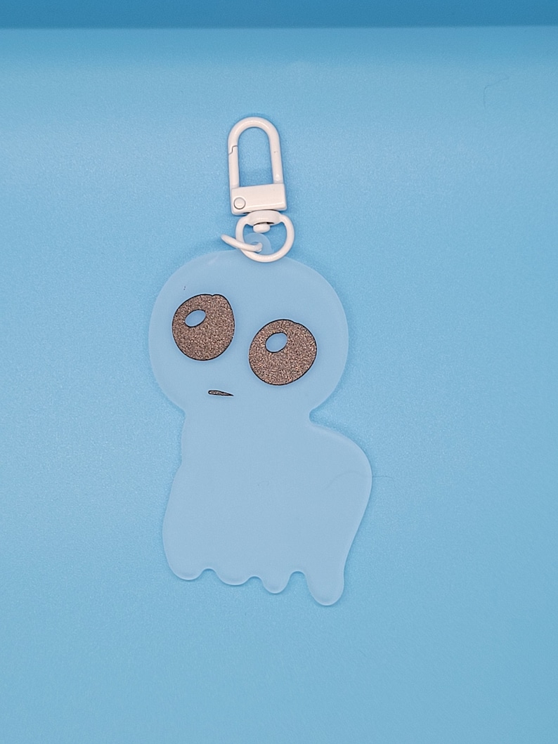 May include: A light blue ghost keychain with a white metal ring. The ghost has two black eyes and a small mouth.