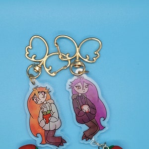 May include: Two cartoon character keychains with gold metal key rings. The characters are a girl with orange hair holding a red strawberry and a girl with purple hair holding a red apple. Each keychain has a red strawberry charm attached.